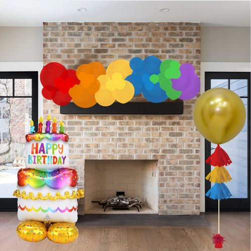 Colorful birthday decorations including balloons and a cake-shaped balloon in front of a brick fireplace.